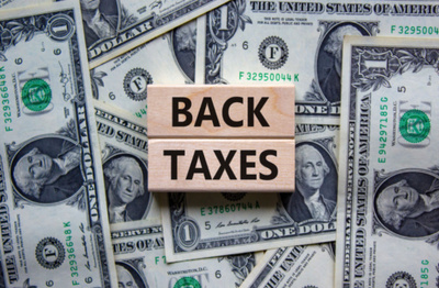 An individual, business, and back taxes concept is illustrated using wooden blocks with 'back taxes' written on a nice backdrop of dollar bills.