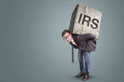 A businessman is crouching beneath a large stone inscribed with the letters IRS to express Las Vegas Tax Liens concept.