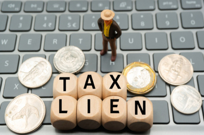 A male figurine is standing on a miniature computer with coins on the keyboard and ‘tax lien’ wooden blocks to illustrate an idea of an individual, business, and back taxes. 